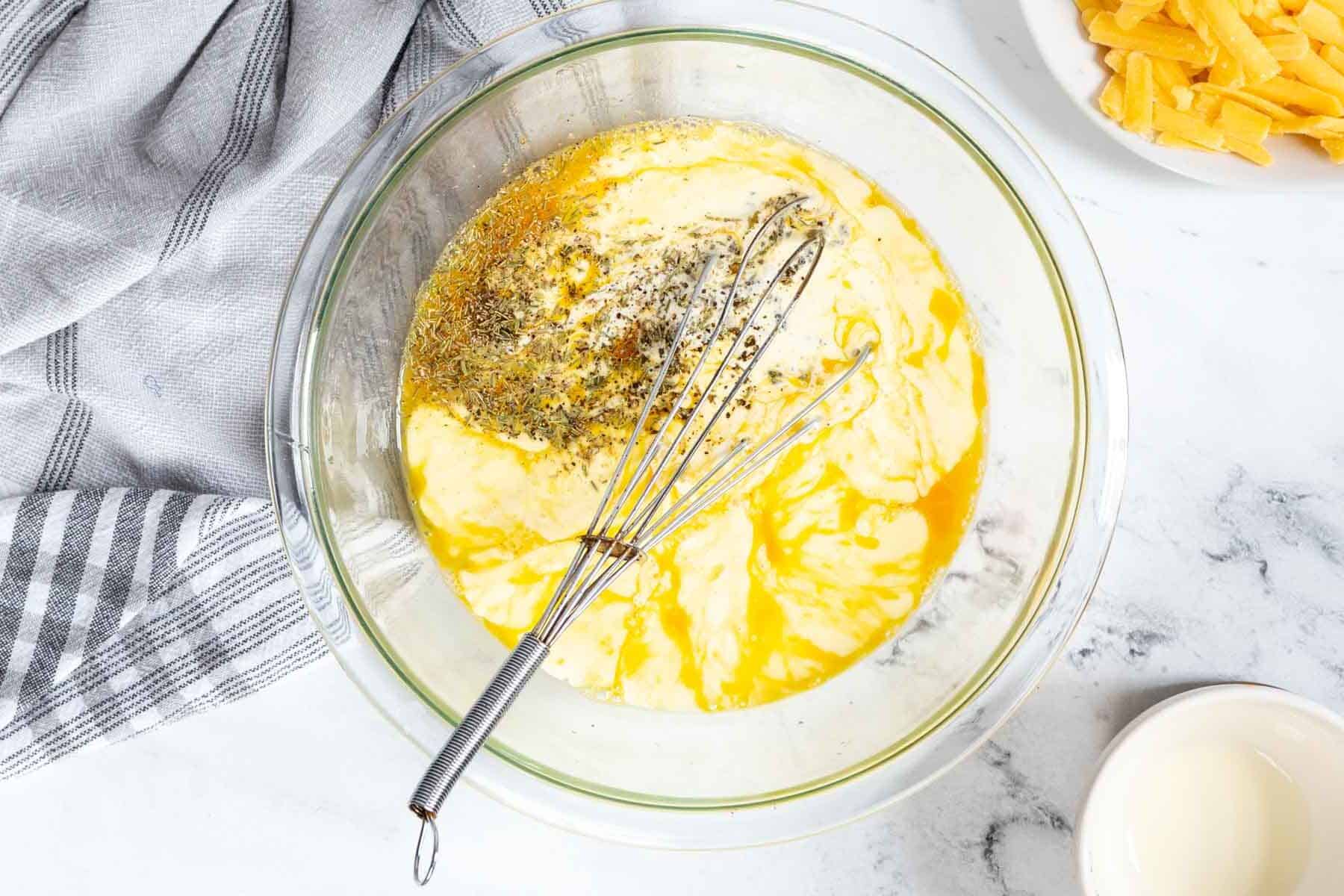 A mixing bowl with eggs and spices sits next to a whisk, striped towel, and sliced cheese on a marble countertop-perfect ingredients for a Ham and Cheese Crustless Quiche.