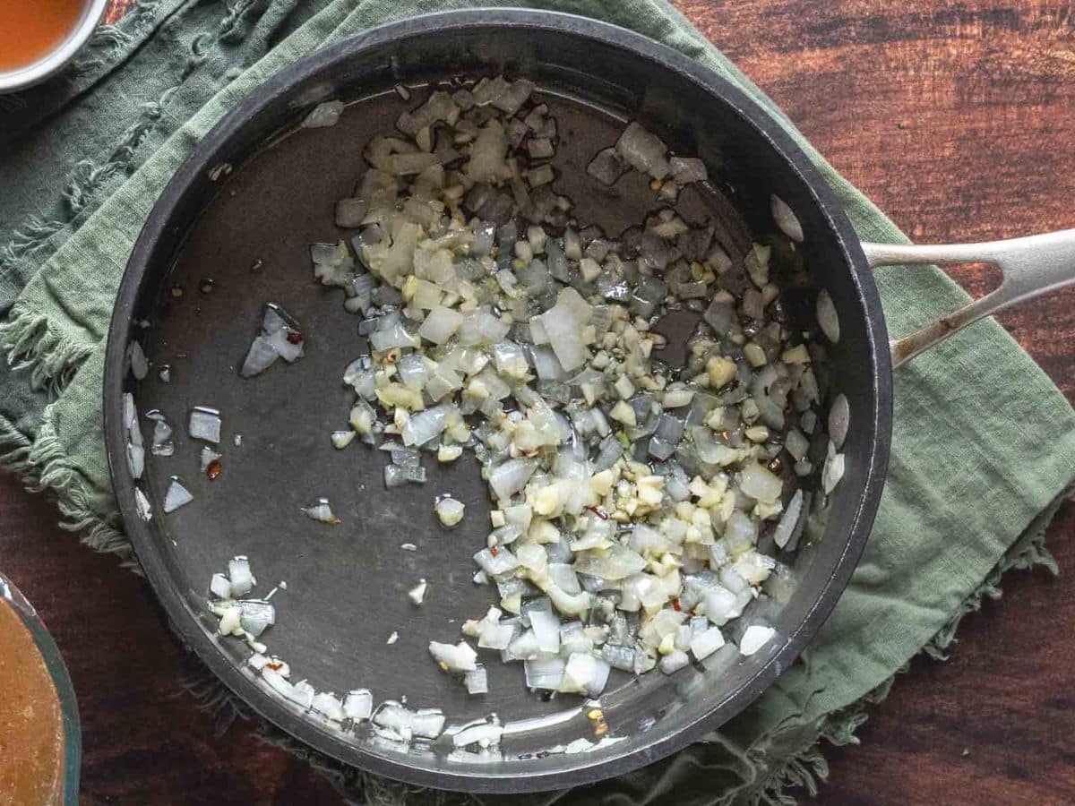 Chopped onions and garlic saut&eacute;ing in a dark pan, ready to infuse their rich aroma into the chimichurri rice, all set on a green cloth atop a wooden surface.