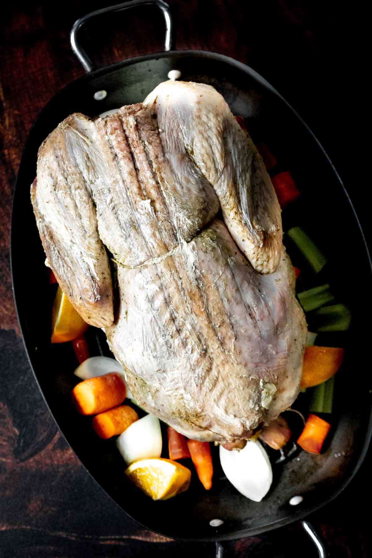 A raw whole turkey, ready for roasting, rests on chopped onions, carrots, celery, and lemon slices in a black pan-a perfect setup if you're learning how to dry brine a turkey for juicy, flavorful results.