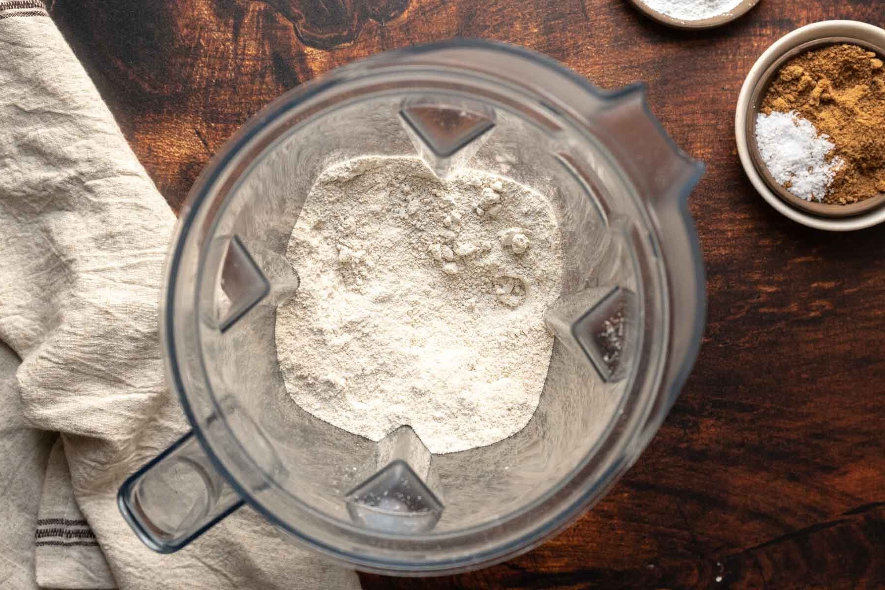 A top-down view of a blender with flour inside, perfect for making oat flour waffles, placed on a wooden surface next to small bowls of spices and salt, alongside a beige cloth.
