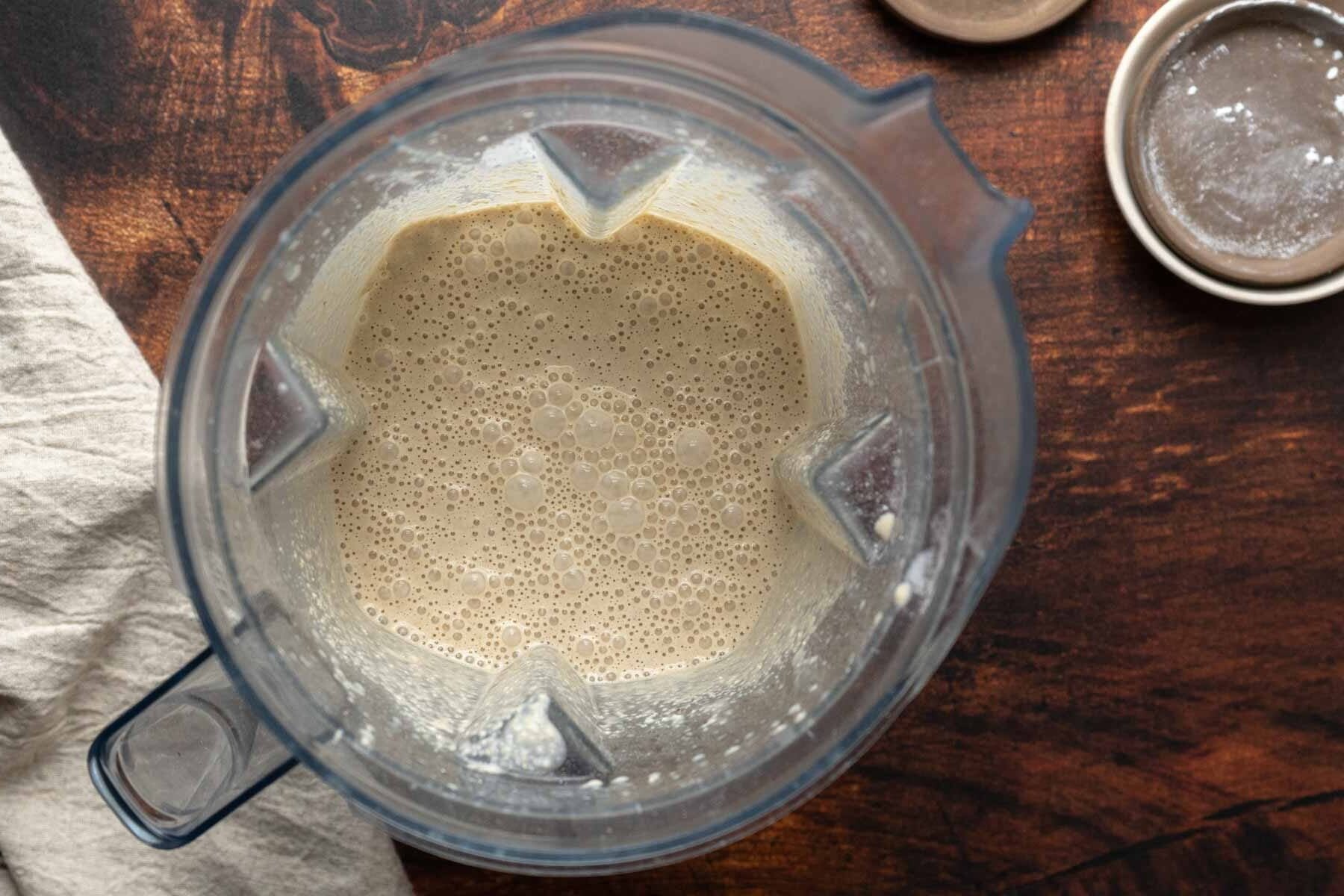 A blender filled with a frothy, beige liquid-perfect for making oat flour waffles-sits on a wooden surface next to a lid and a cloth.