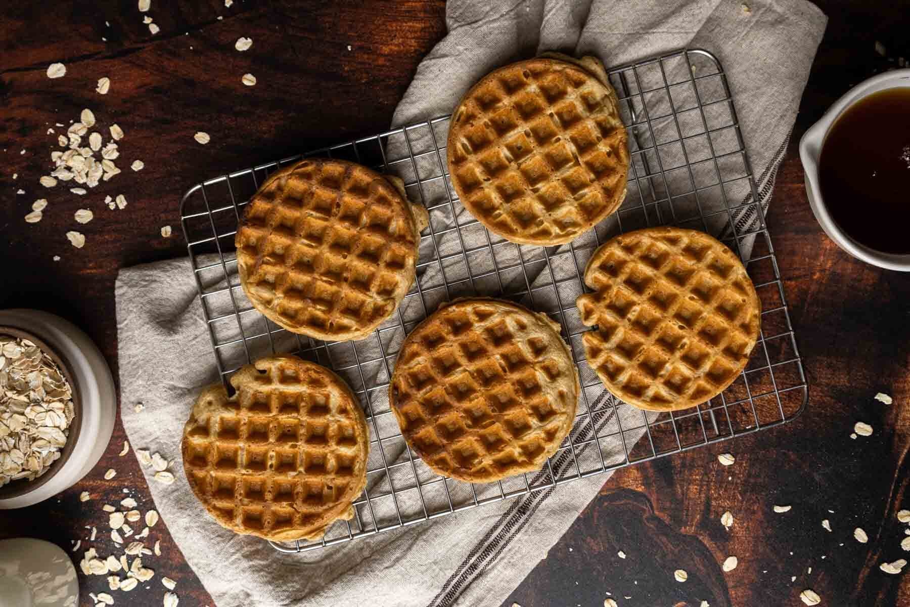 Five round oat flour waffles are cooling on a metal rack over a beige cloth, with a cup of syrup and a bowl of oats nearby on a wooden surface.