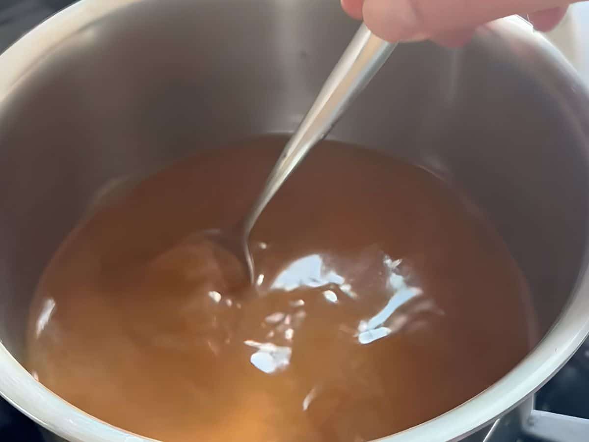 A hand stirs a pot of turkey gravy with a metal spoon on a stovetop, preparing rich gravy for a roast turkey.