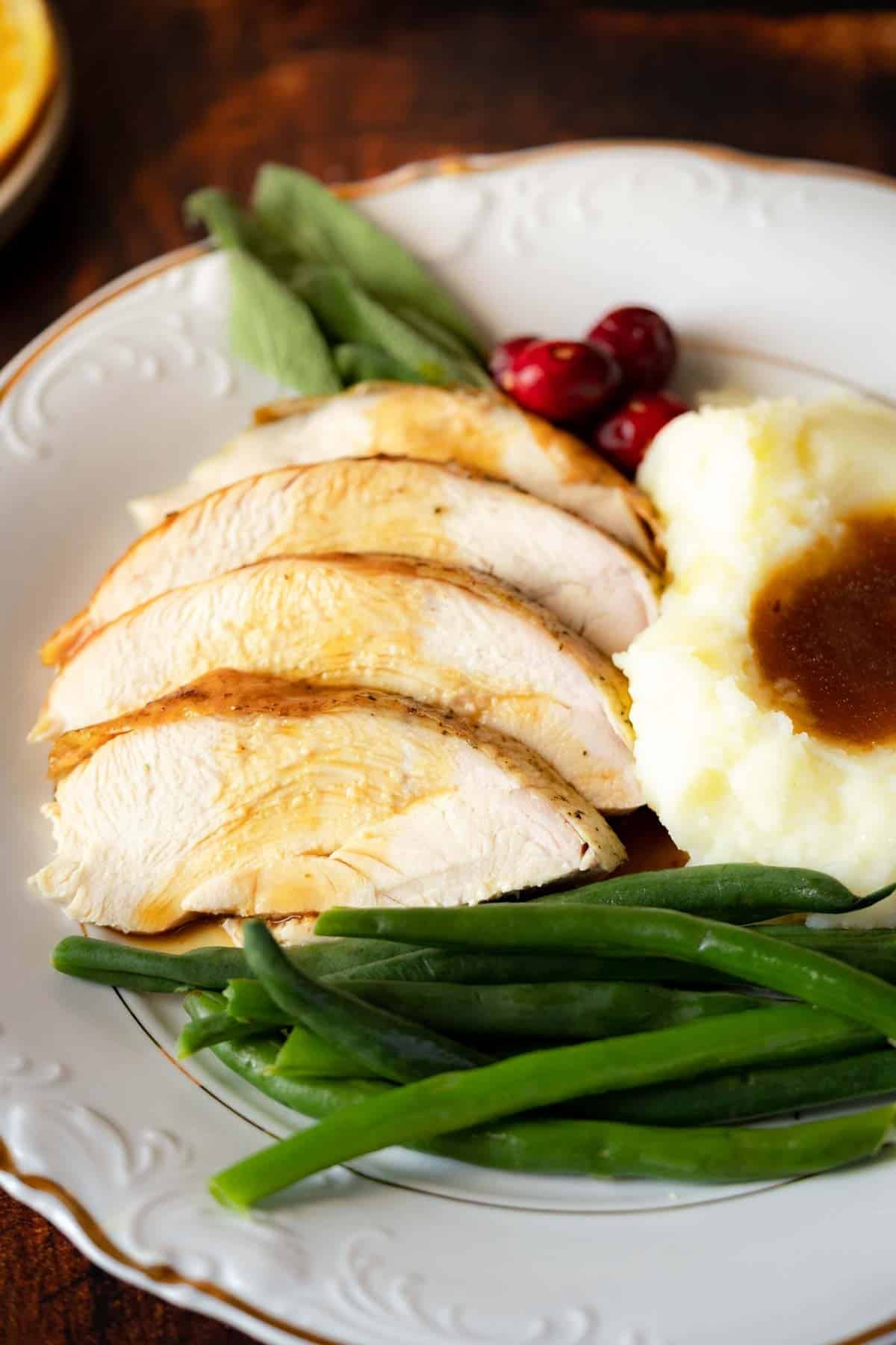 Sliced roast turkey breast with mashed potatoes and gravy, green beans, cranberries, and sage leaves on a white plate.