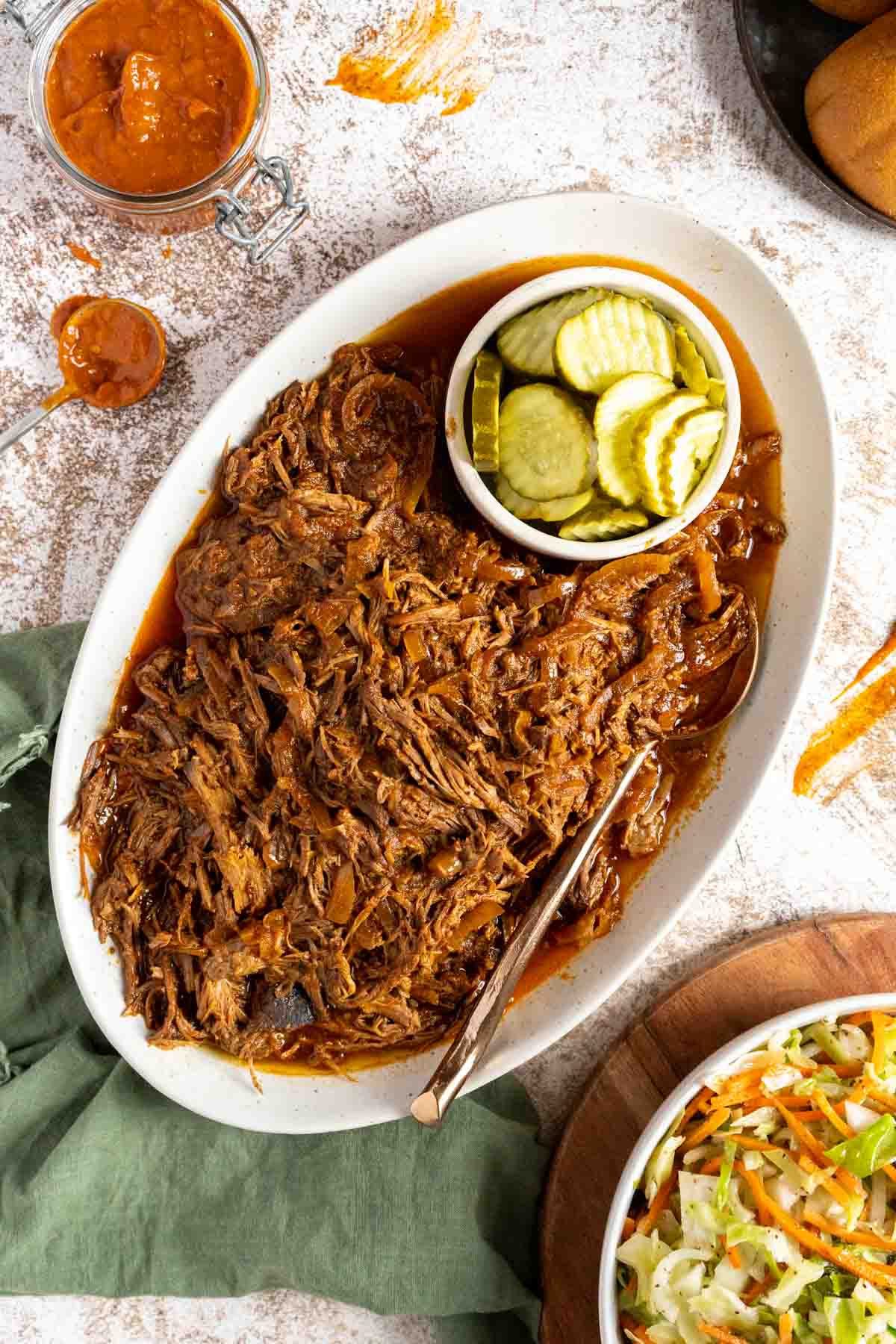 BBQ shredded beef on a serving platter with pickles and a side of coleslaw.