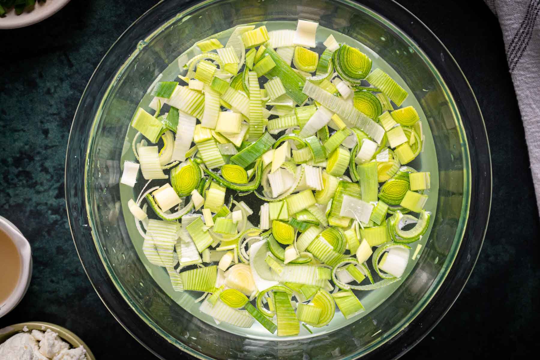A glass bowl filled with chopped leeks soaking in water, surrounded by ingredients for Broccoli Asparagus Soup, with other food items partially visible around the bowl.