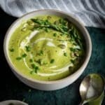 A bowl of broccoli asparagus soup garnished with asparagus tips, pumpkin seeds, chopped chives, and a swirl of cream sits next to a spoon on a dark surface.