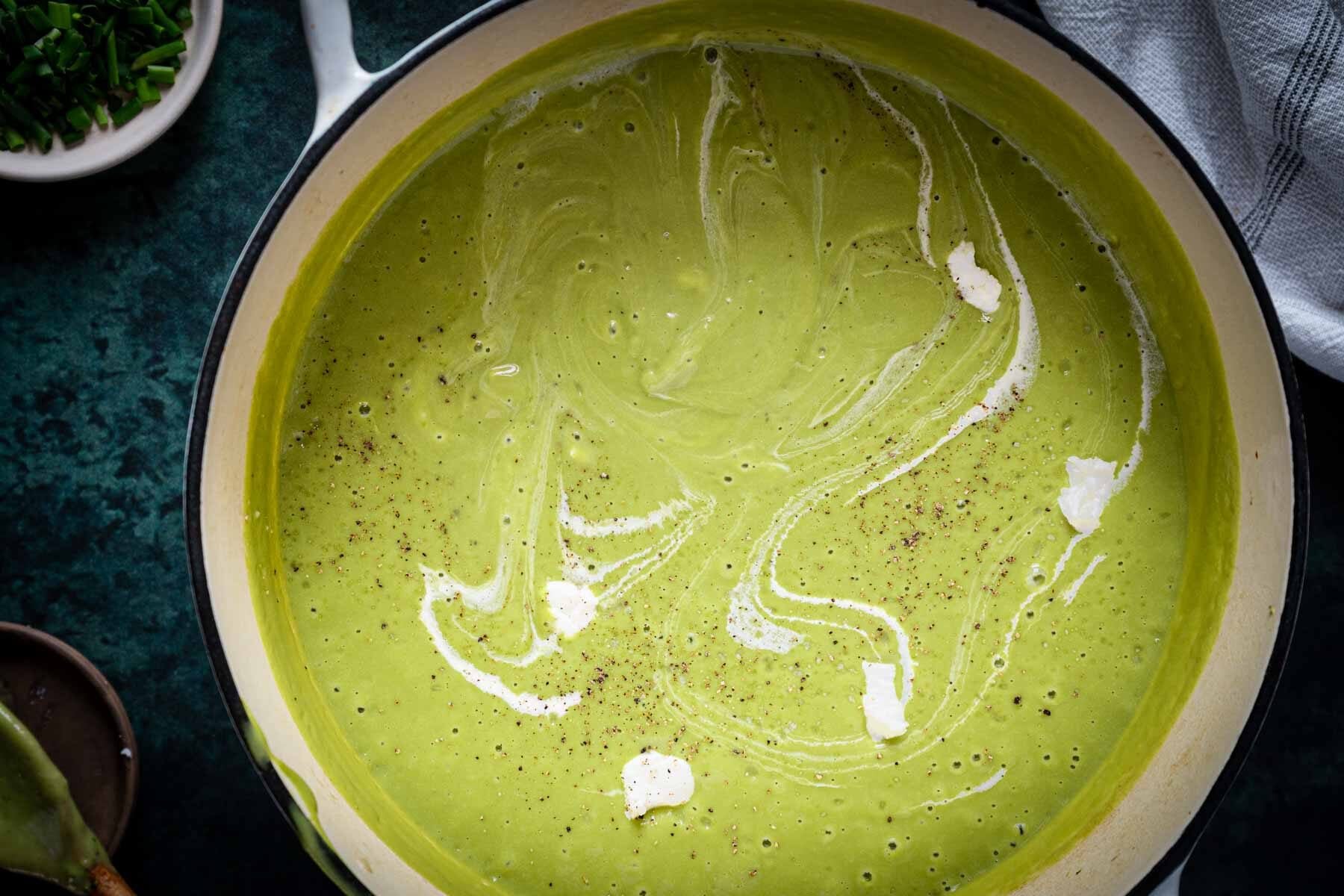 A pot of creamy green Broccoli Asparagus Soup with white swirls and a sprinkle of black pepper on top.