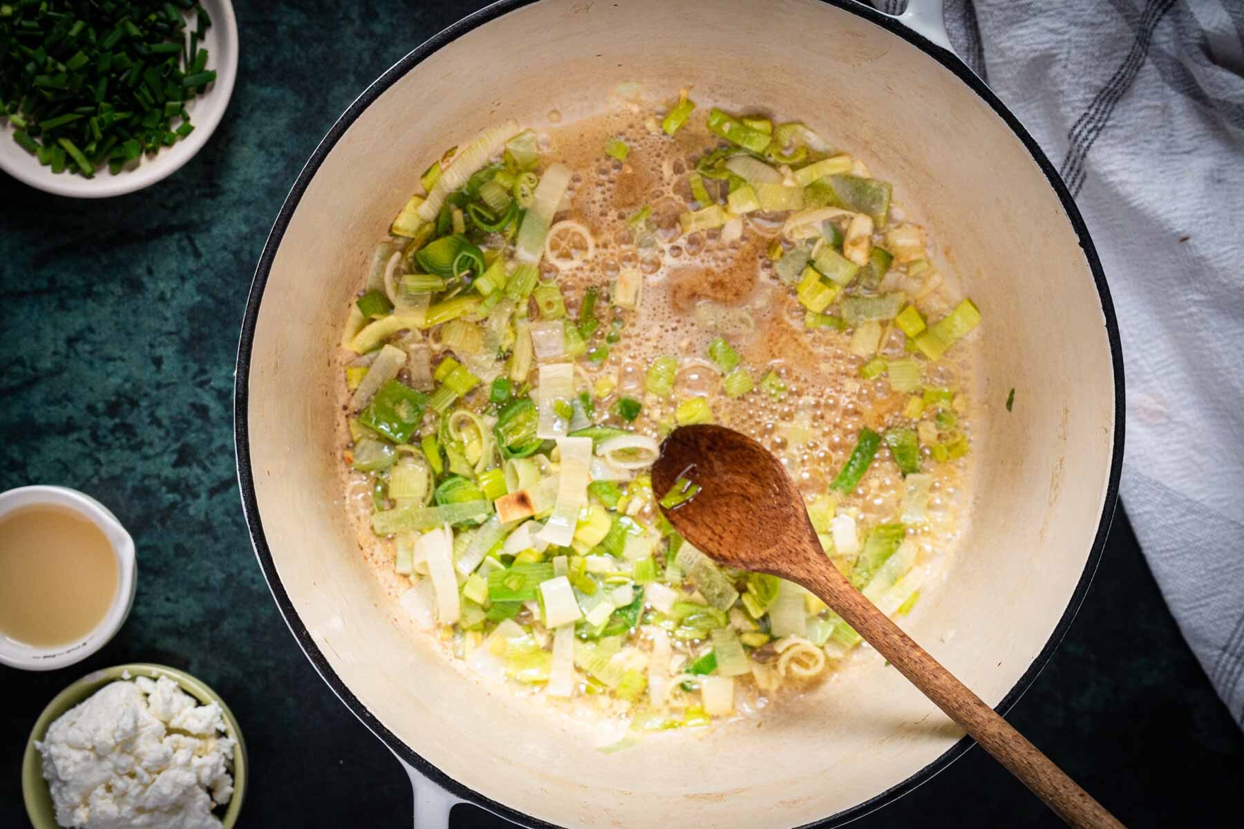 Chopped leeks saut&eacute;ing in a white pot with a wooden spoon, surrounded by small bowls of ingredients-perfect beginnings for a flavorful Broccoli Asparagus Soup-set on a dark countertop.