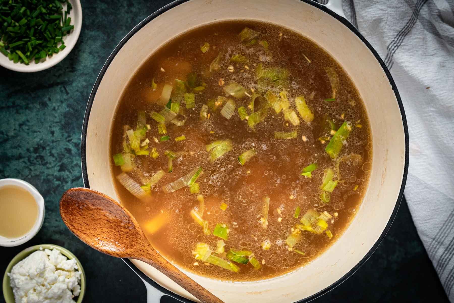 A pot of vegetable broth with chopped leeks and herbs, perfect for making Broccoli Asparagus Soup, shown with a wooden spoon and ingredient bowls nearby on a dark countertop.