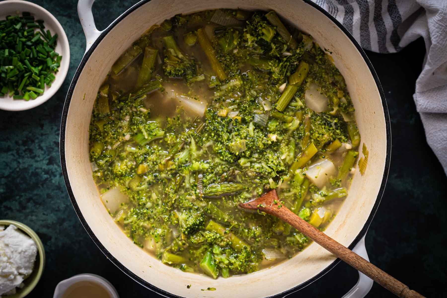 A white pot filled with Broccoli Asparagus Soup, with a wooden spoon resting inside. Bowls of chives and a white ingredient sit nearby on a dark surface.