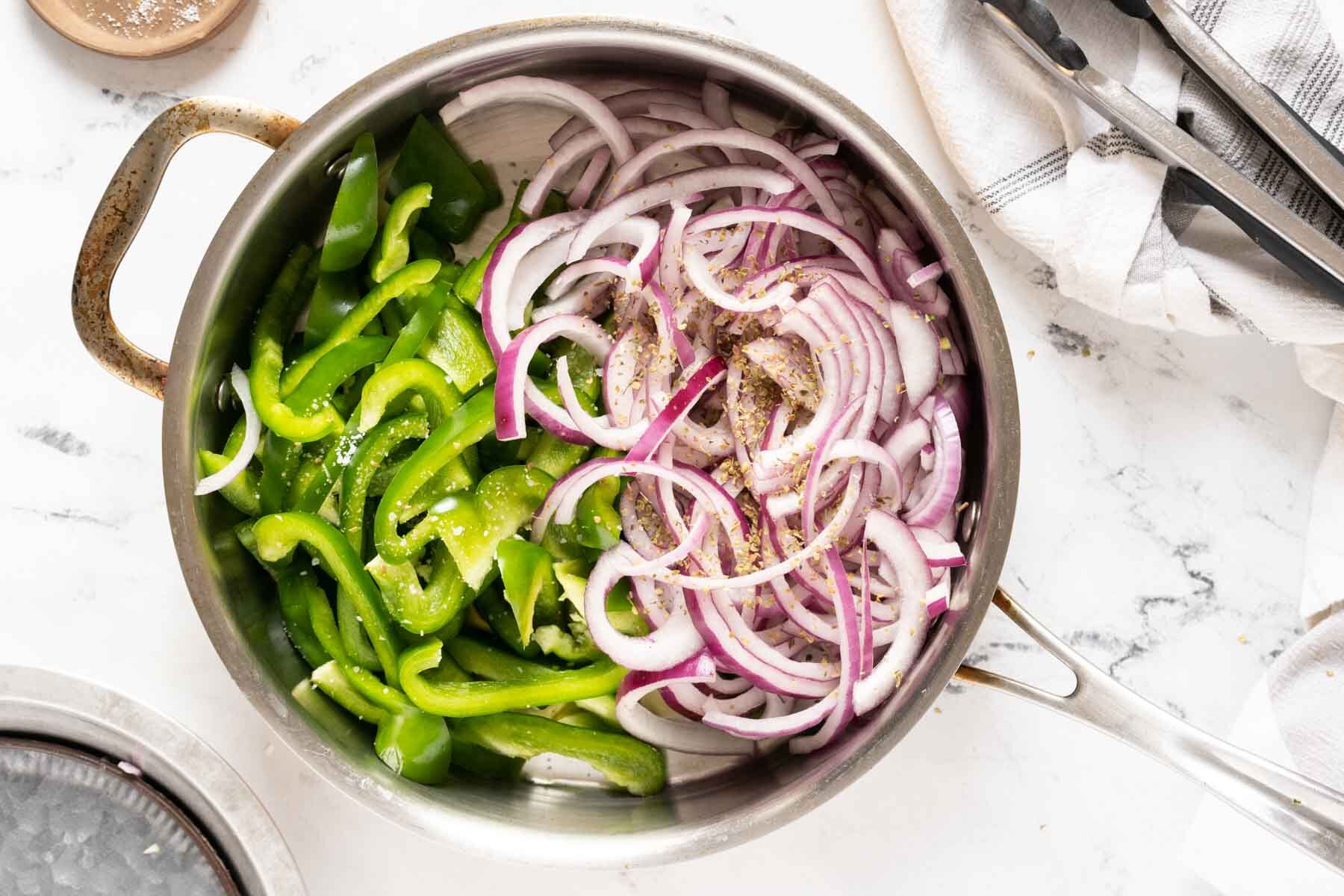 A stainless steel pan containing sliced green bell peppers and red onions, seasoned with black pepper, sits on a white marble countertop next to a striped towel and metal tongs-perfect for making Chipotle-style fajita veggies.