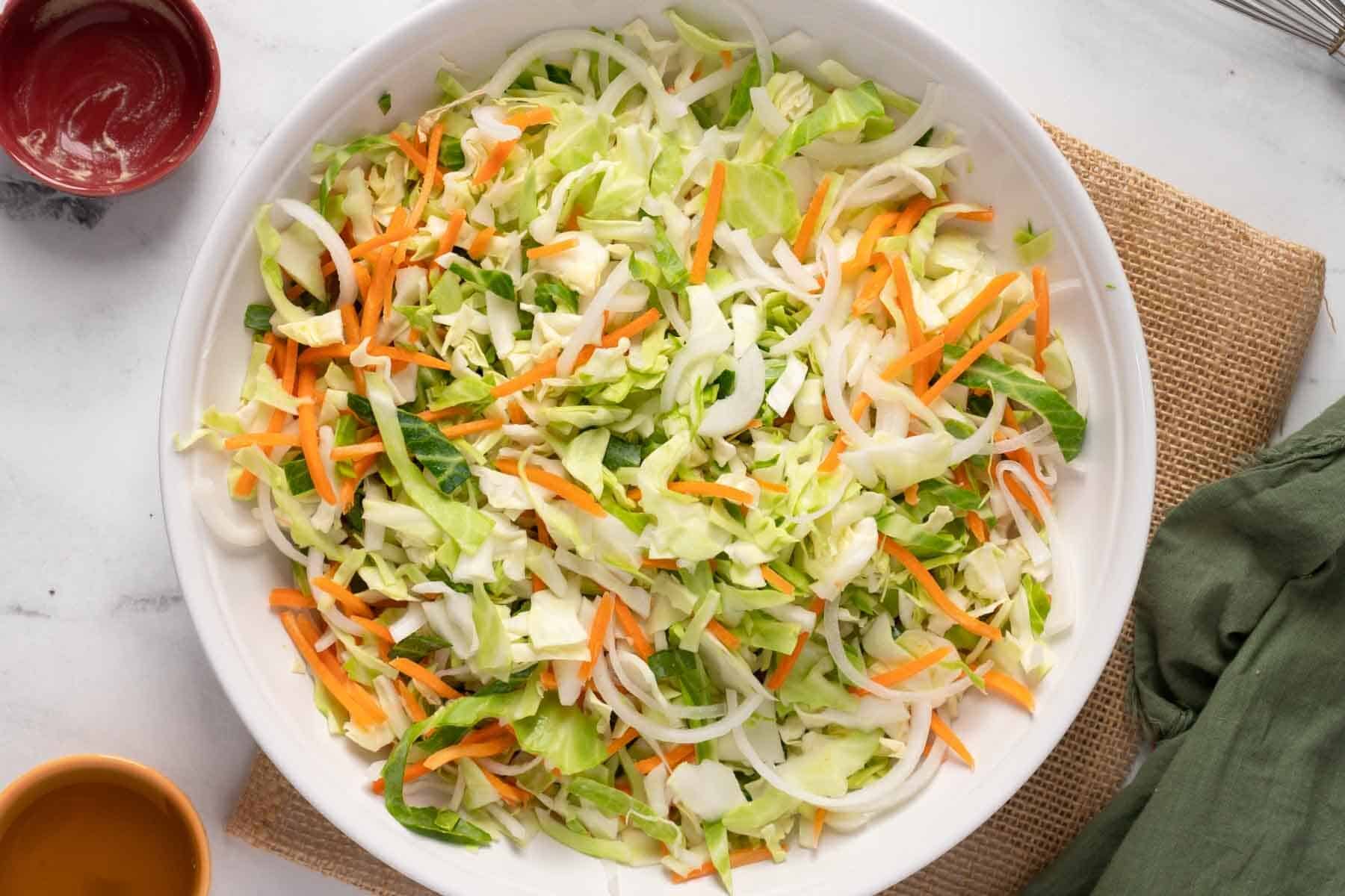 A bowl filled with chopped cabbage, carrots, and onions, sits on a table with a green napkin and small dishes nearby.
