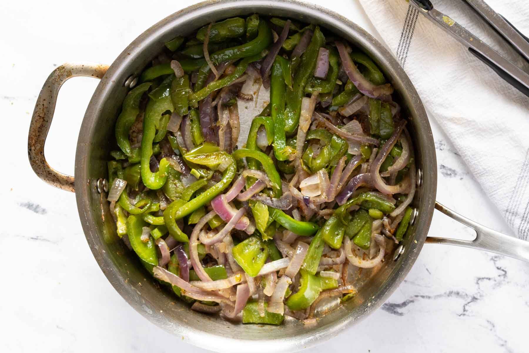 A stainless steel pan on a white surface contains sizzling Chipotle-style fajita veggies, with saut&eacute;ed green bell peppers and red onions. A pair of metal tongs rests on a white cloth nearby.