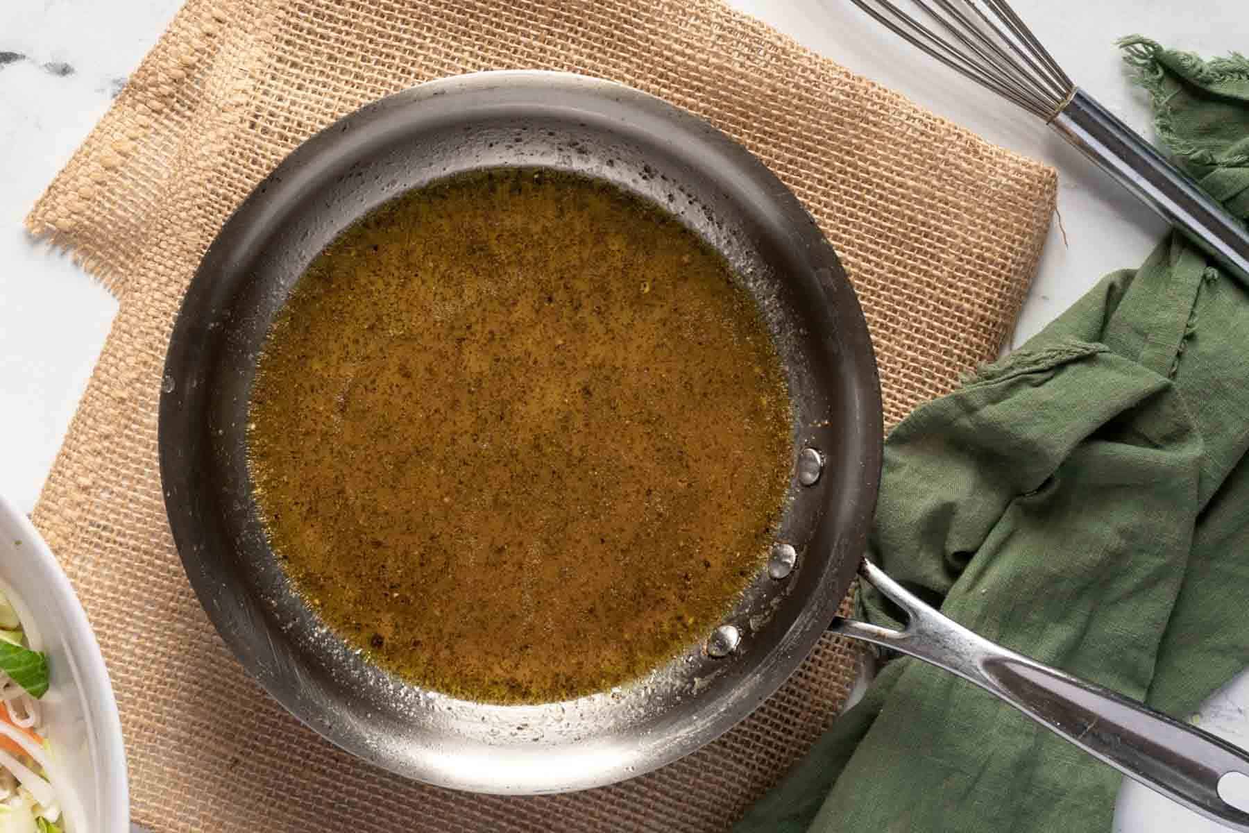 A stainless steel pan with vinegar dressing for Old Fashioned Coleslaw with Vinegar sits on a burlap placemat, next to a green cloth napkin and a metal whisk.