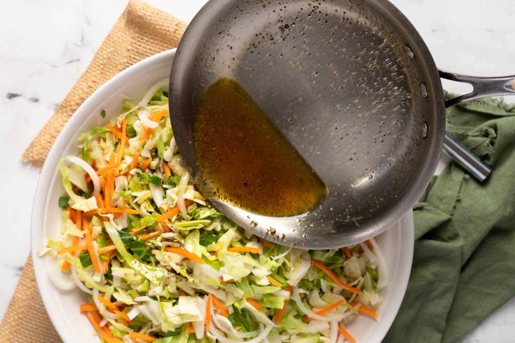 A metal pan pours dressing over a bowl of shredded cabbage, carrots, and green onions for an Old Fashioned Coleslaw with Vinegar, set on a table with a green napkin and brown placemat.