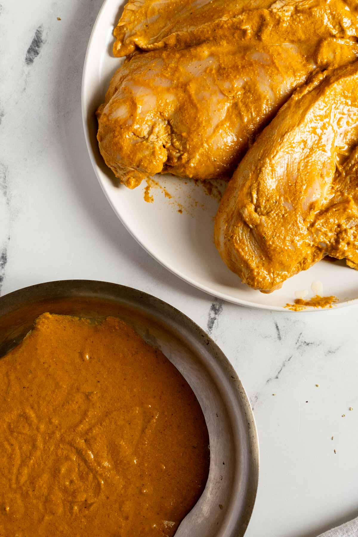 Raw chicken breasts coated in orange marinade on a white plate, with extra marinade in a metal bowl on a white marble surface.