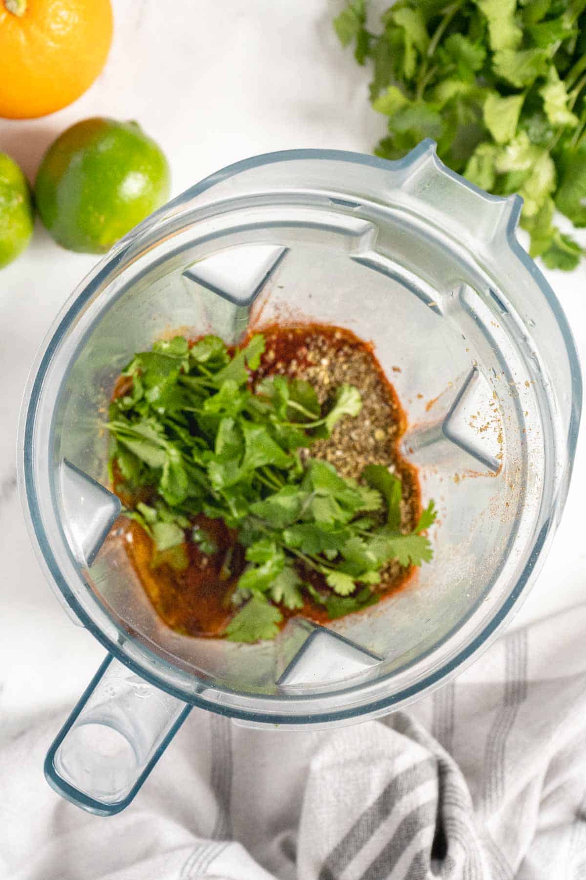 A blender containing fresh cilantro, spices, and liquids on a white countertop with limes, an orange, and cilantro in the background.