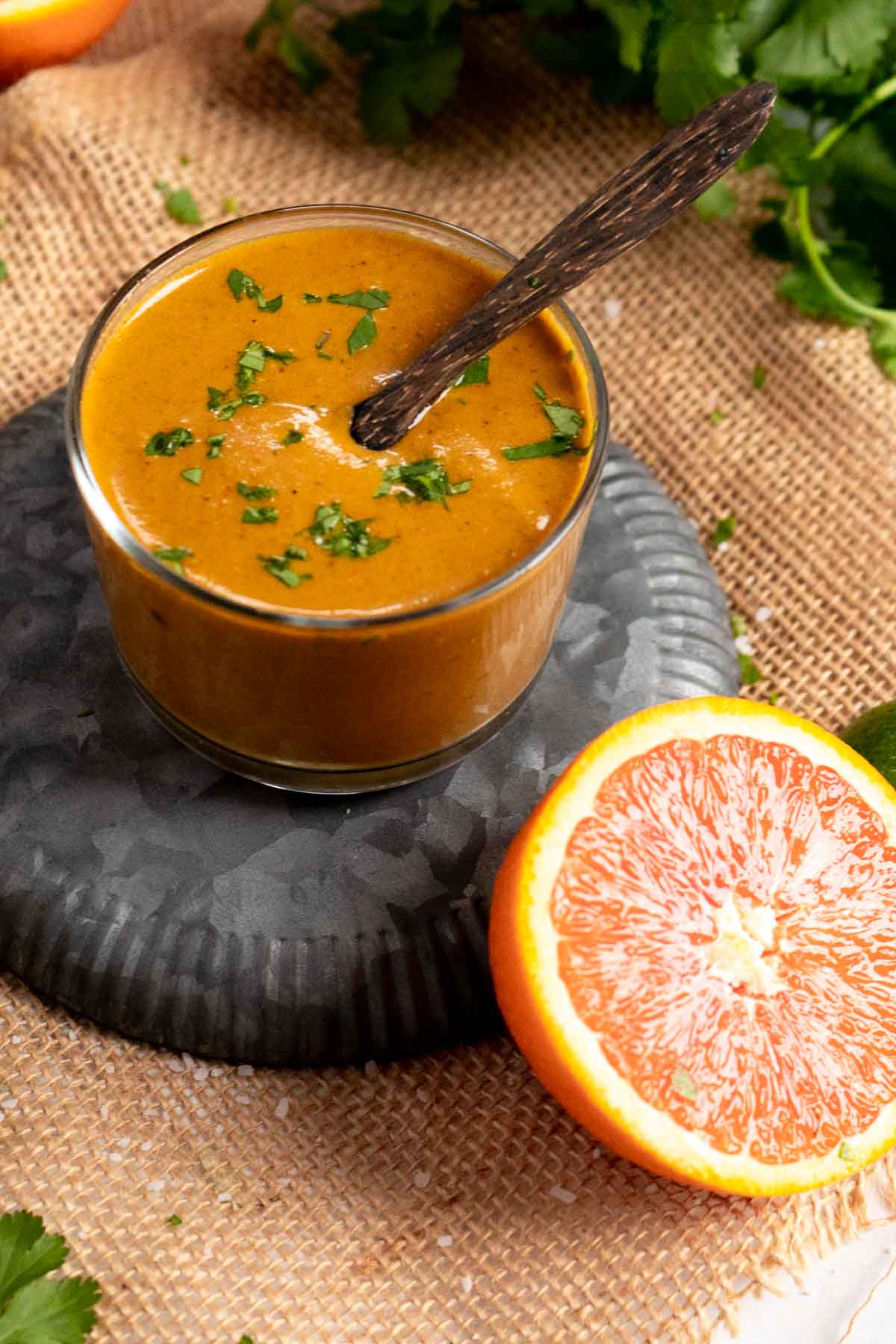 A glass cup filled with pollo asado marinade garnished with chopped herbs, with a spoon inside, placed on a dark plate beside a halved orange and fresh greens.
