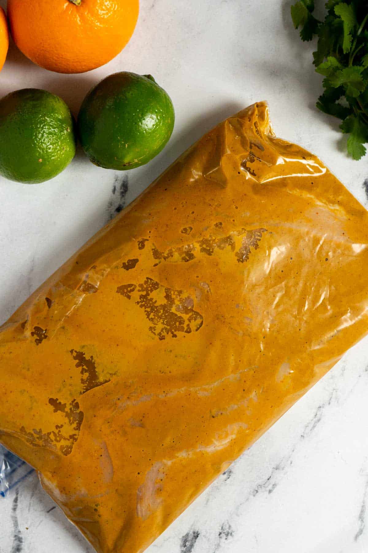 A sealed plastic bag filled with orange marinade sits on a marble surface near fresh oranges, limes, and cilantro.