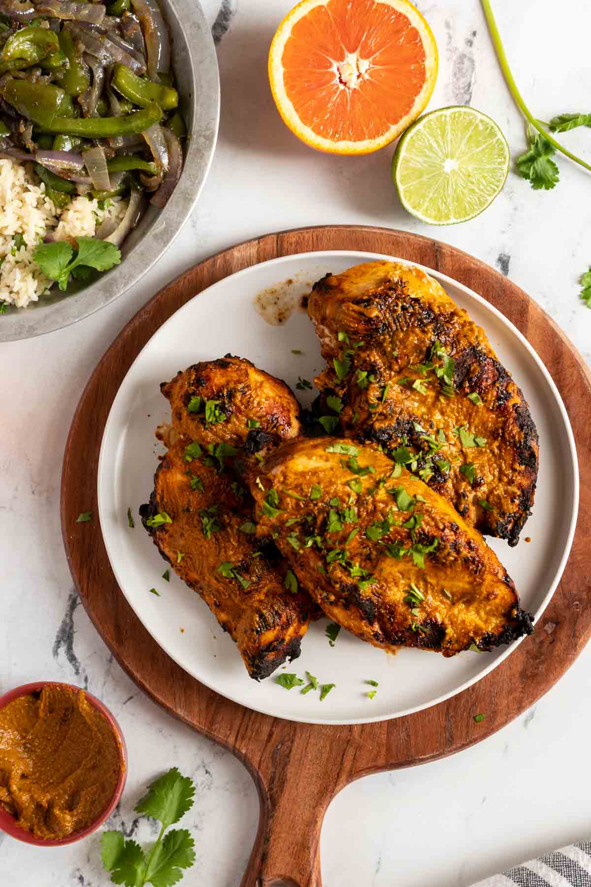 Grilled chicken breasts garnished with chopped cilantro on a white plate, with a side of sauteed vegetables, rice, an orange half, and lime half.