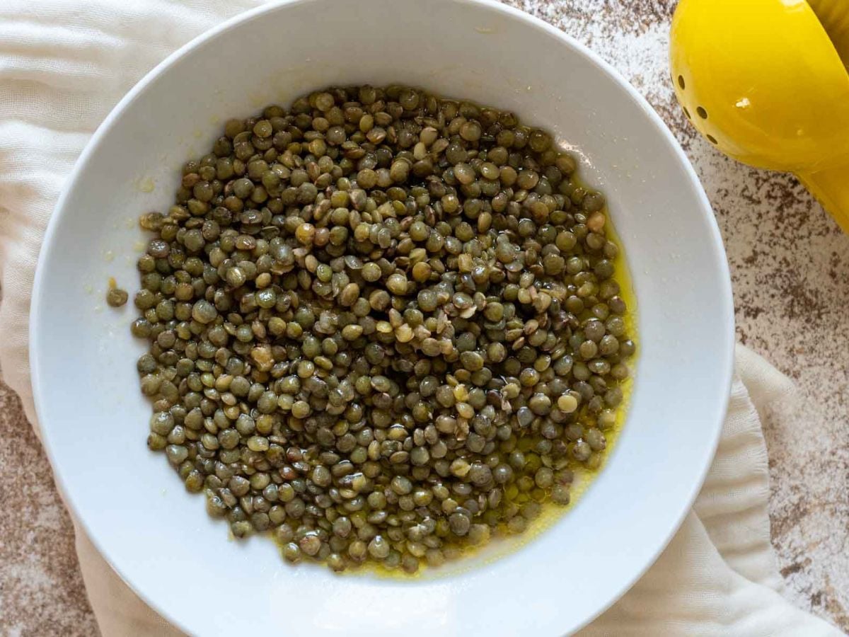 A white bowl filled with cooked green lentils, perfect for a lentil tabbouleh, sits in a light, oily liquid on a beige cloth with a yellow citrus juicer partially visible in the top right corner.