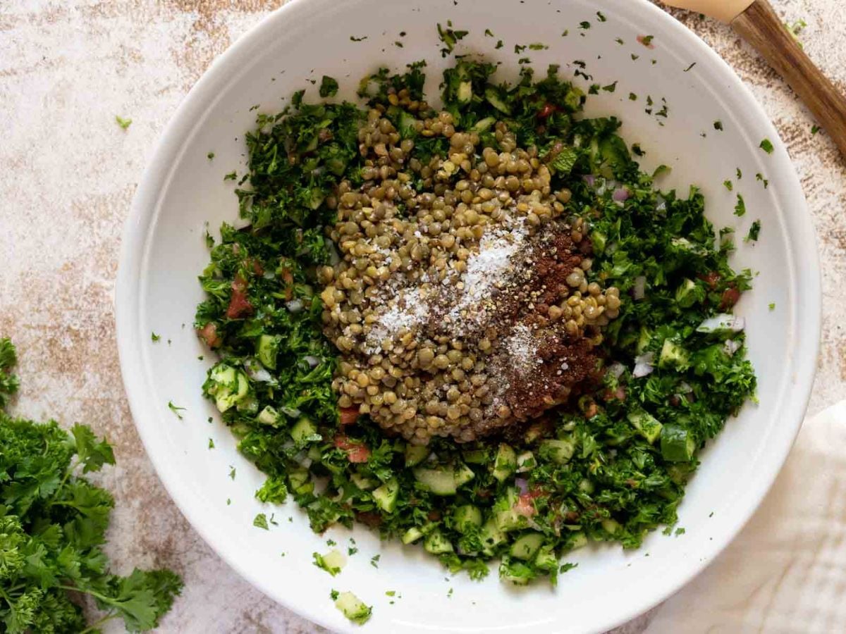 A white bowl filled with lentil tabbouleh-chopped parsley, diced cucumbers, tomatoes, cooked lentils, and seasoning powders-rests on a light-colored surface. A bunch of fresh parsley and a wooden spoon are nearby.