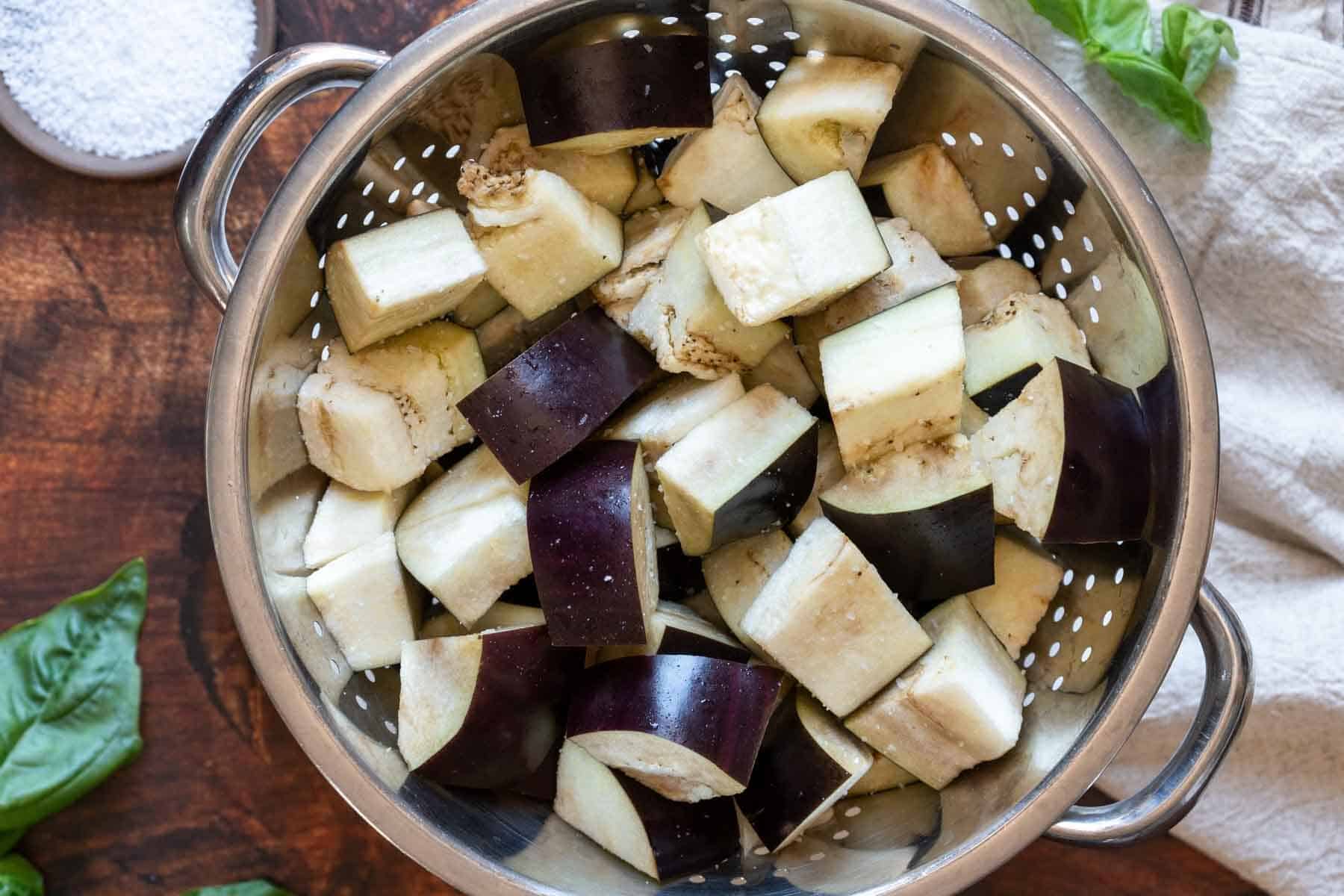 Cubed pieces of raw eggplant, perfect for making eggplant caponata, sit in a metal colander on a wooden surface with fresh basil leaves and a white cloth nearby.