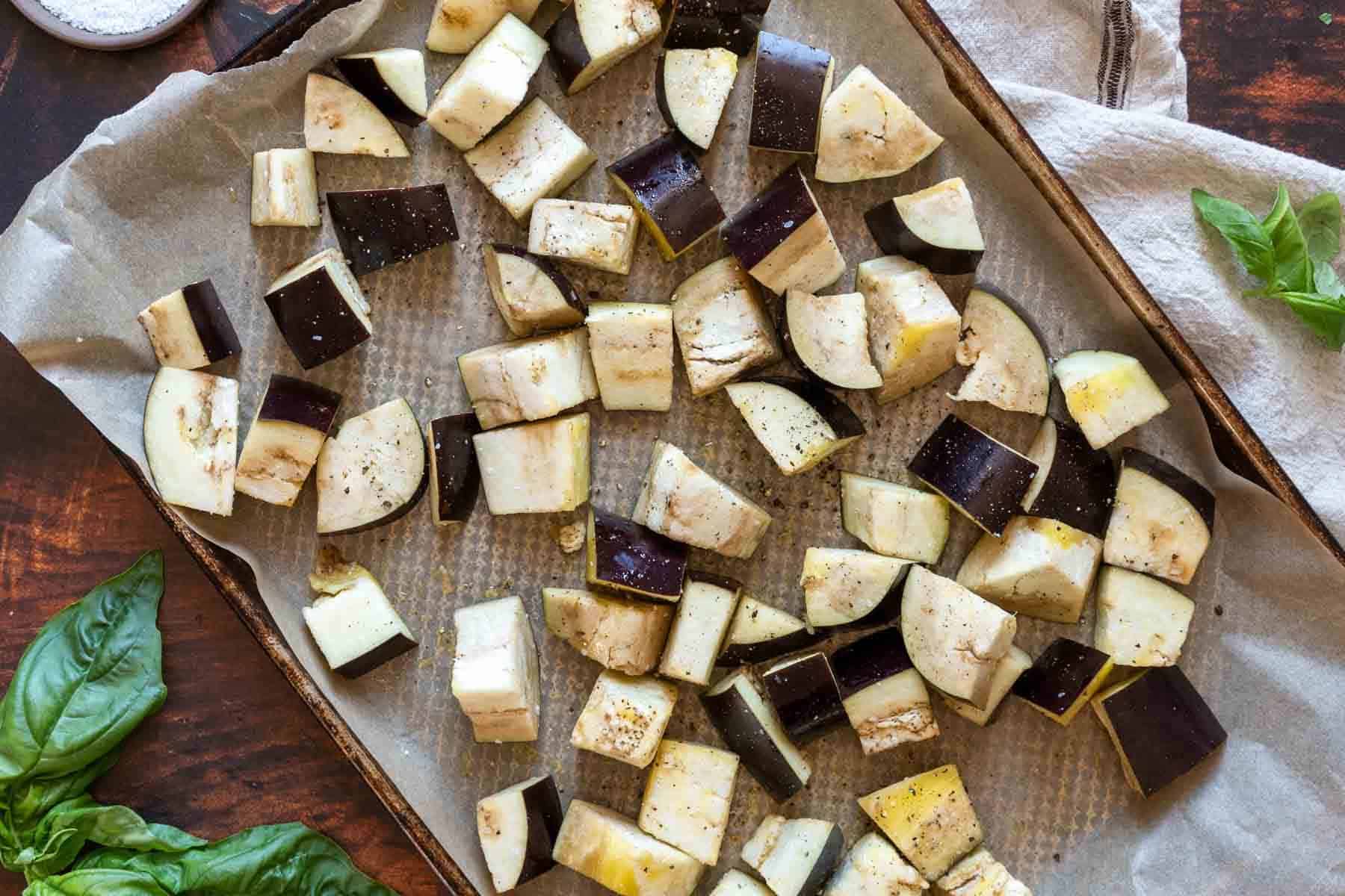 Chunks of raw eggplant, perfect for eggplant caponata, are spread out on a parchment-lined baking sheet, seasoned with salt and pepper, ready for roasting. Fresh basil leaves are visible nearby.