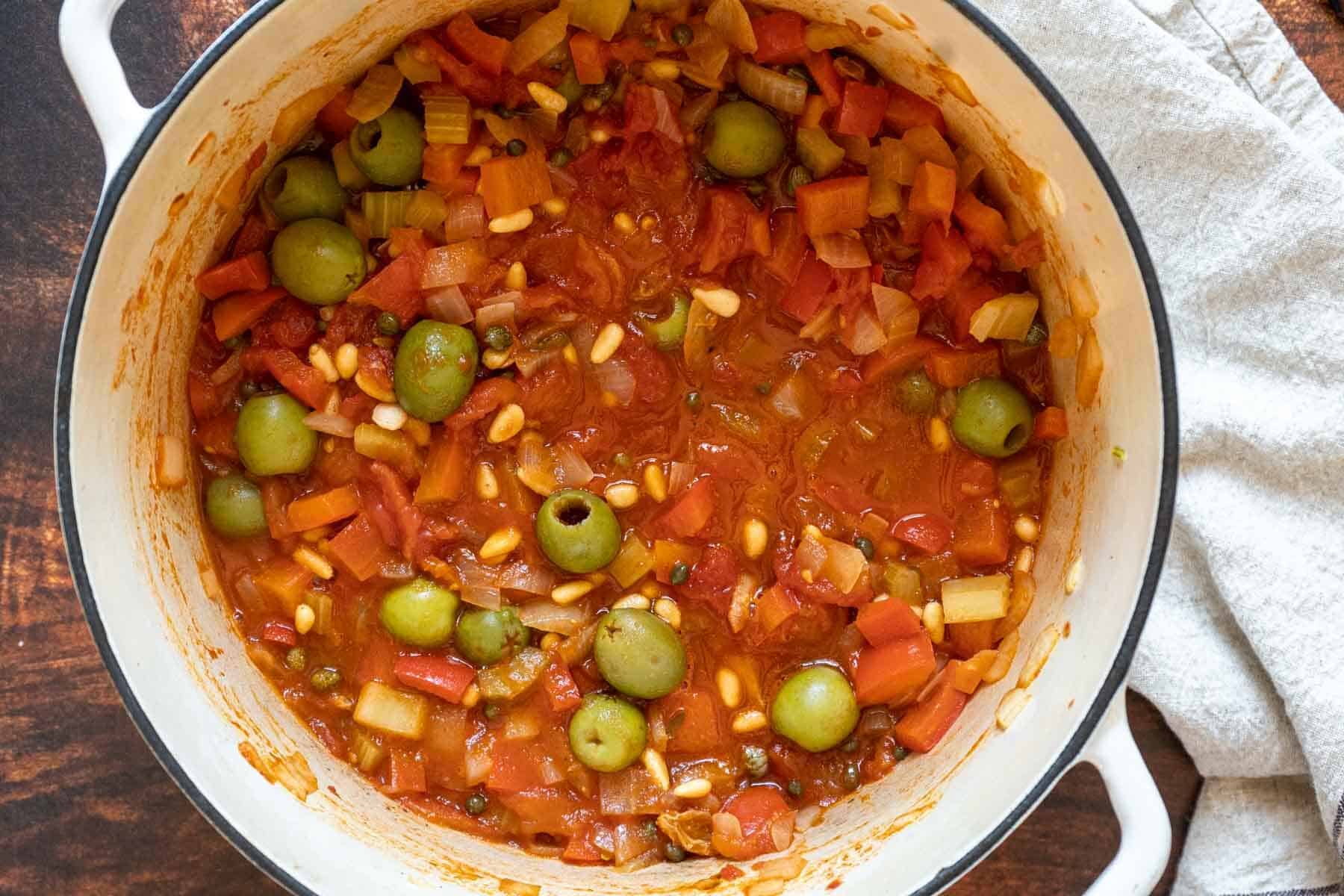 A white pot filled with eggplant caponata, a tomato-based stew featuring green olives, pine nuts, chopped vegetables, and capers, sits on a wooden surface next to a beige cloth.