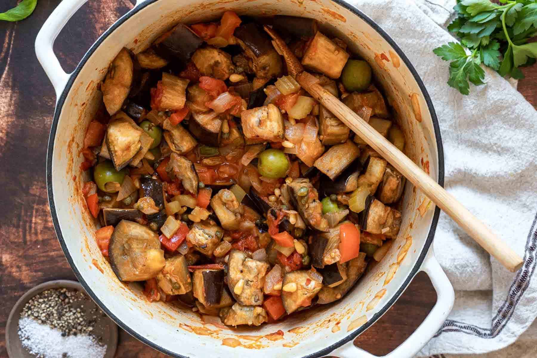A white pot filled with a savory vegetable stew inspired by eggplant caponata, featuring eggplant, tomatoes, olives, and onions, with a wooden spoon resting inside.