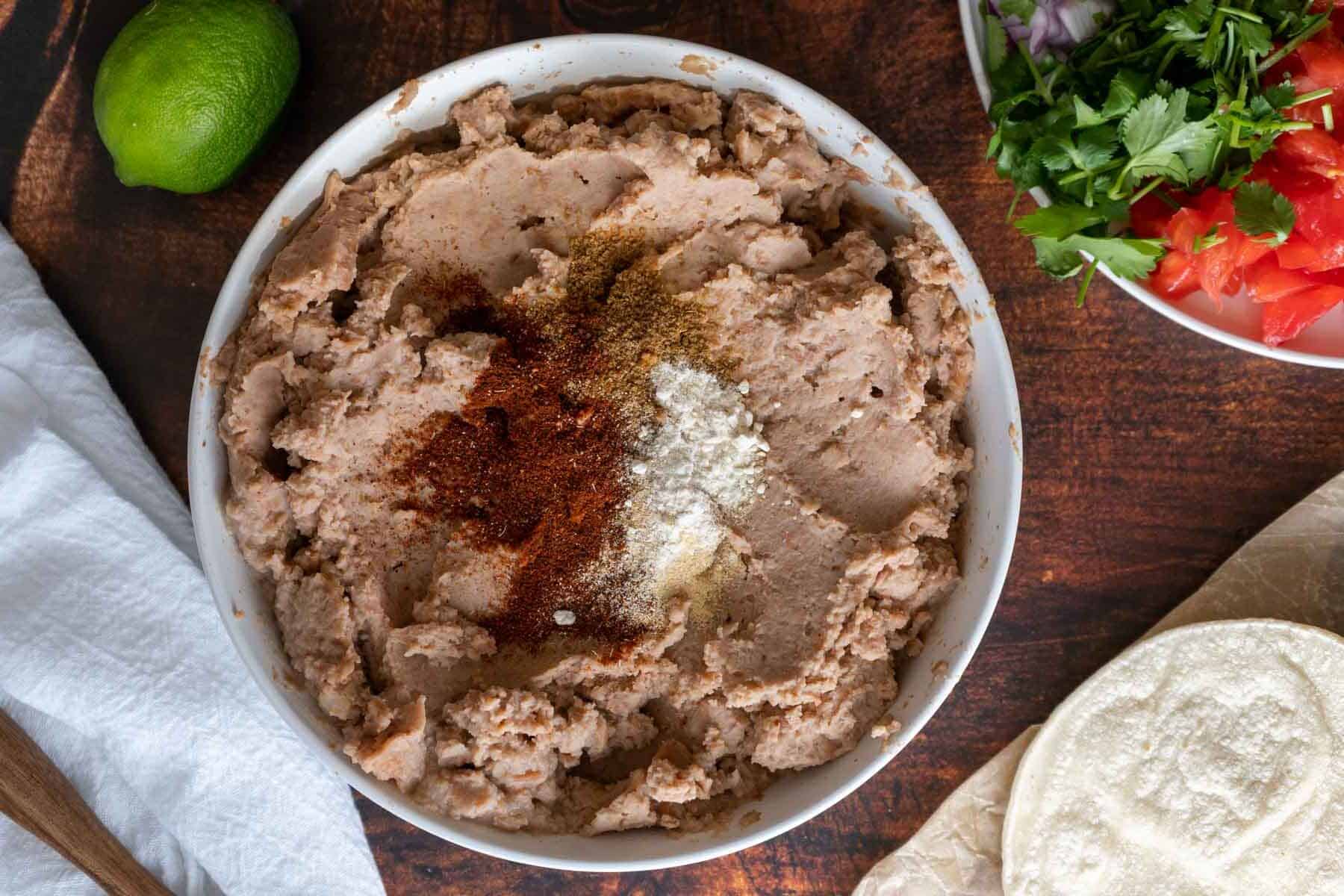 A bowl of refried beans with spices, paired perfectly for making bean and cheese tacos, surrounded by a lime, a white napkin, a plate of diced vegetables, and tortillas on a wooden surface.