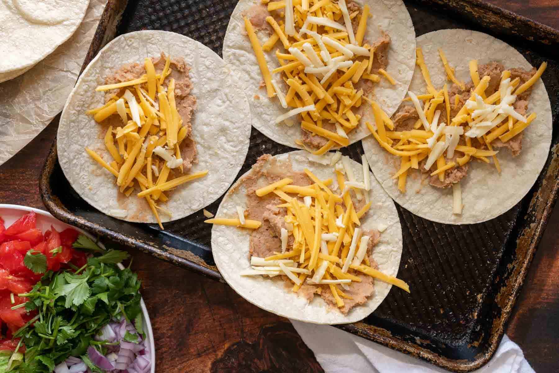 Four corn tortillas topped with refried beans and shredded cheese rest on a baking sheet, ready to become savory bean and cheese tacos. Nearby are fresh garnishes of chopped tomatoes, cilantro, and onions.
