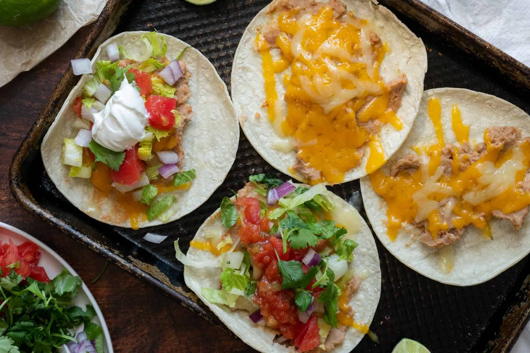 A baking sheet with four bean and cheese tacos topped with a medley of ingredients, including crisp lettuce, ripe tomatoes, tangy onions, fresh cilantro, and creamy sour cream.
