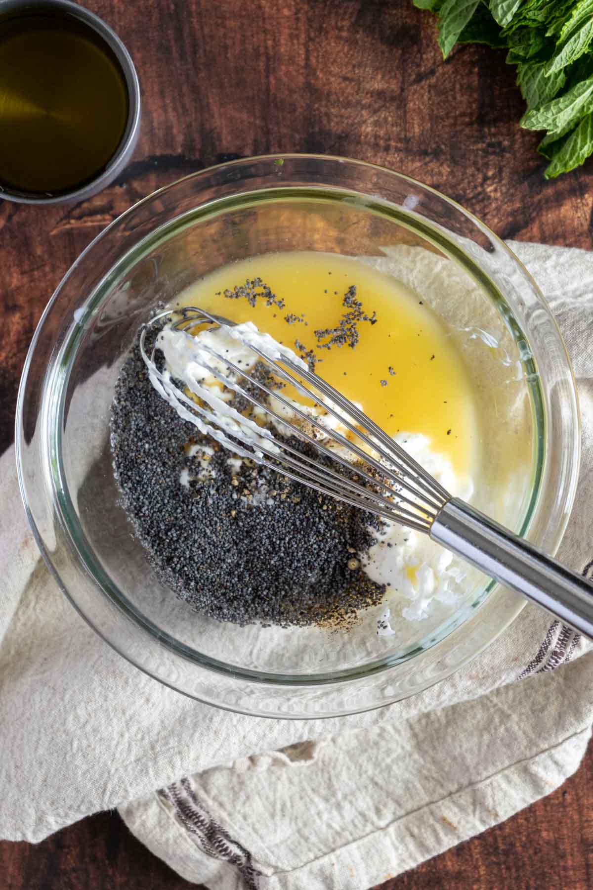 A glass bowl with a metal whisk mixing poppy seeds, creamy white dressing, and yellow liquid on a beige cloth, set on a wooden surface. A cup of oil and leafy greens are visible in the background.