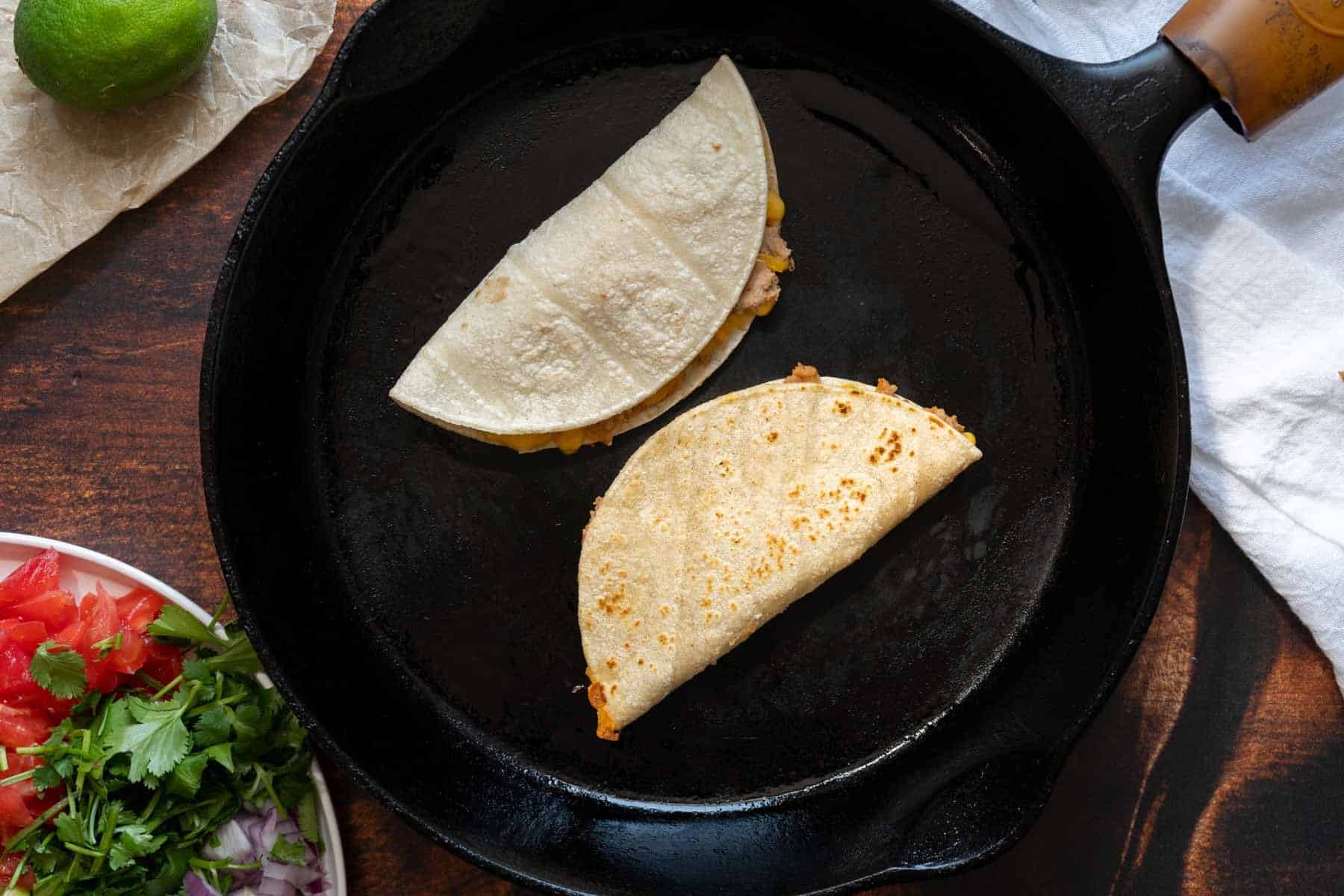 Two folded bean and cheese tacos are sizzling on a black skillet, while fresh ingredients like chopped tomatoes and greens wait nearby.
