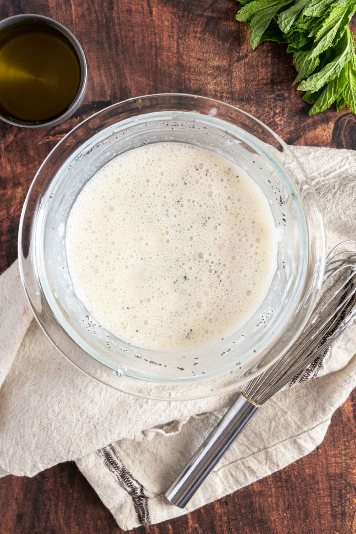 A glass bowl with a frothy white liquid and a whisk inside sits on a beige cloth, next to a small cup of oil and a bunch of fresh mint leaves on a wooden surface.