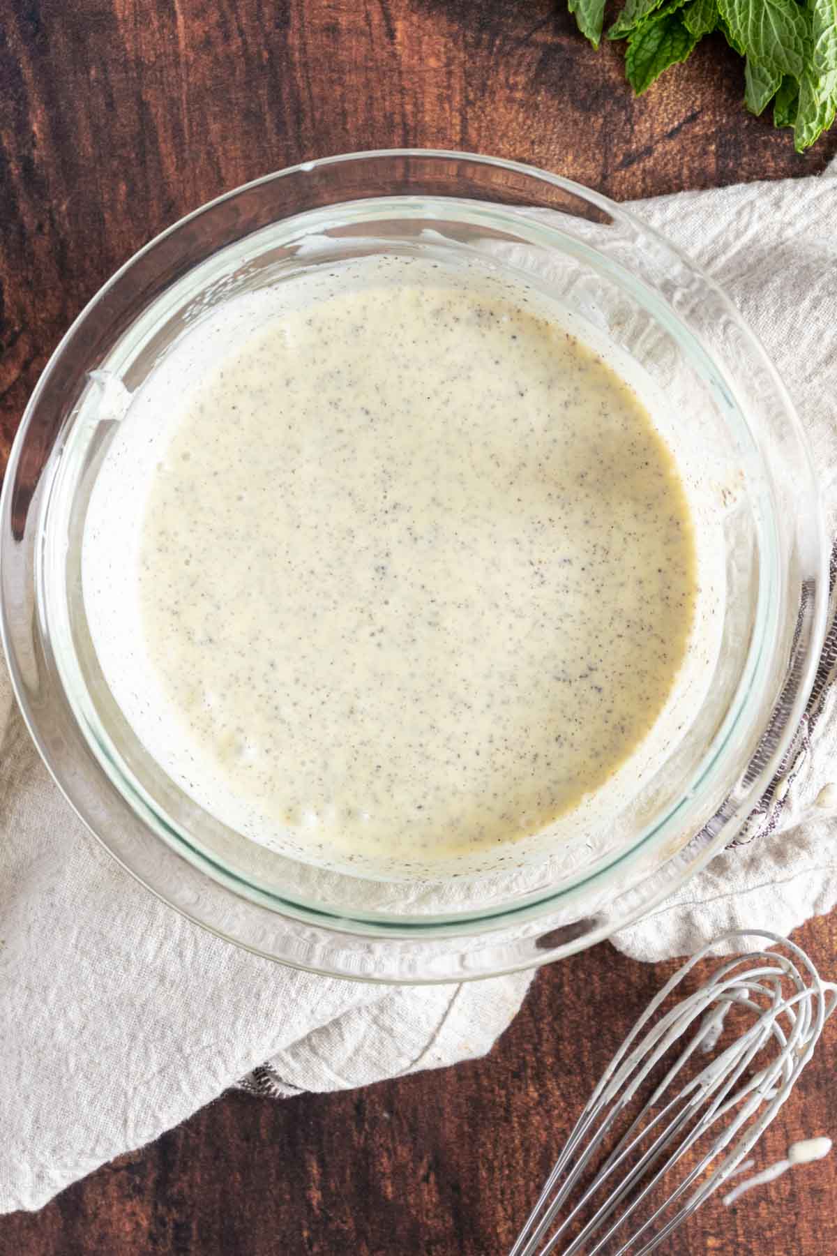 A clear glass bowl containing a creamy, speckled dressing or sauce sits on a white cloth atop a dark wooden surface. A metal whisk and fresh mint leaves are nearby.