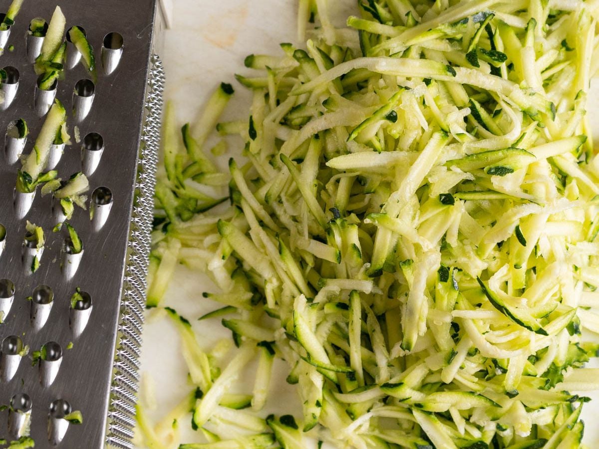 Close-up of a metal box grater next to a mound of freshly grated zucchini on a white surface. Strands of shredded zucchini, perfect for making creamy zucchini risotto, are scattered beside the grater.
