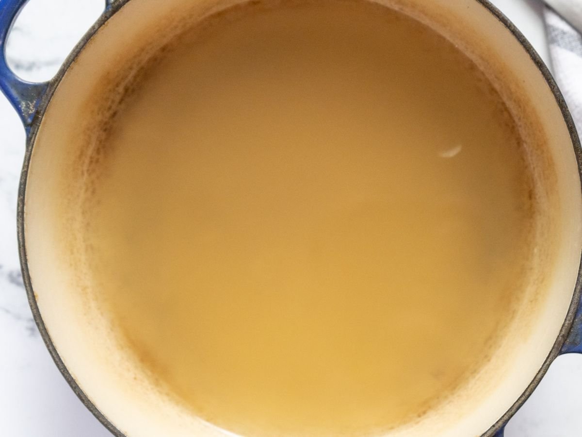 A large pot filled with light brown zucchini risotto broth or stock, with a slight ring of residue around the inner edge. The surface of the liquid is mostly smooth and reflective.