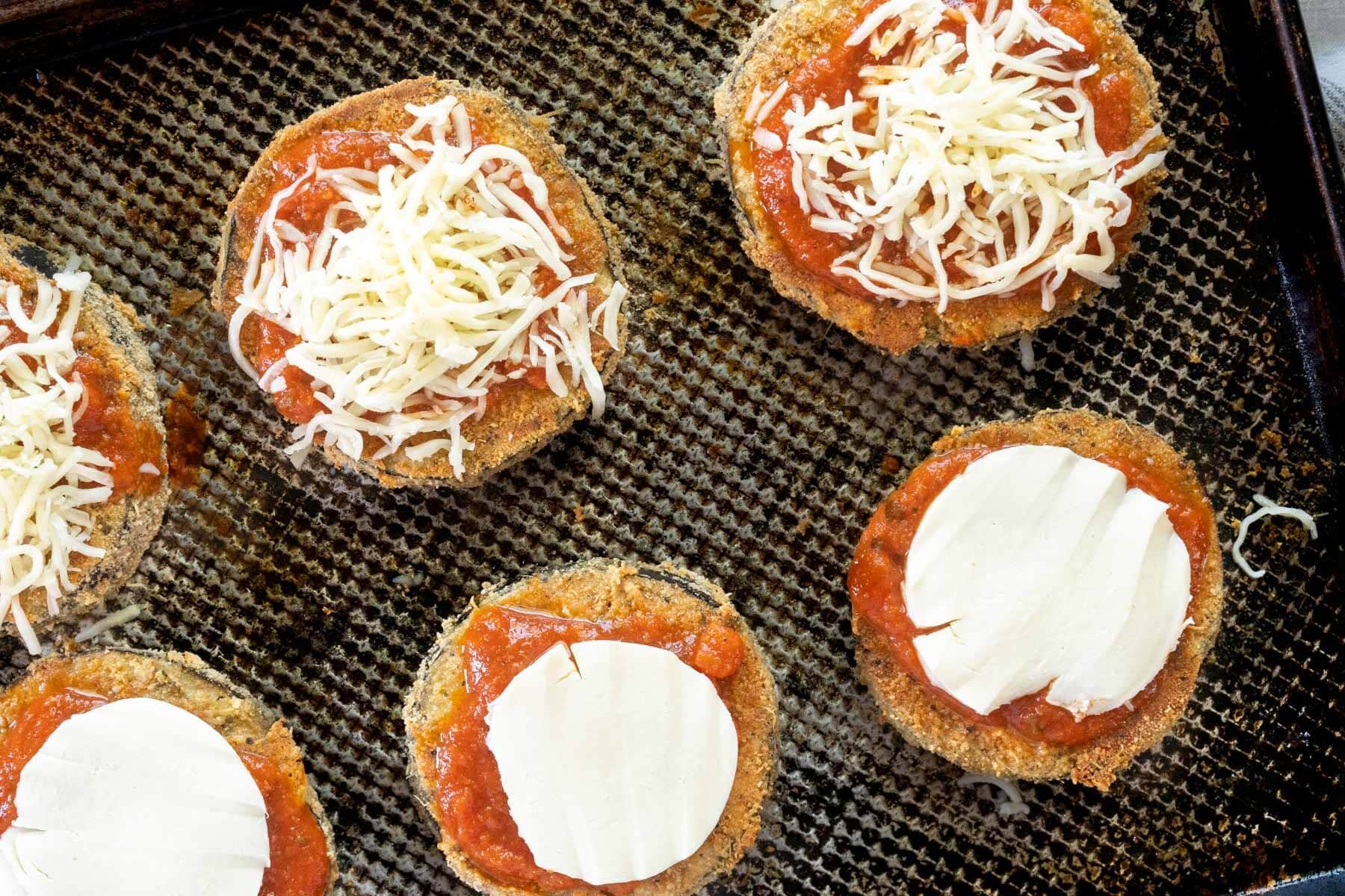Gluten-free eggplant parmesan slices rest on a baking sheet, topped with tomato sauce and mozzarella cheese, ready for the oven.