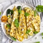 A white plate with grilled zucchini and yellow squash slices, garnished with crumbled cheese and fresh basil leaves, sits beside Huli Huli Chicken on a striped cloth with a grilled lemon half.