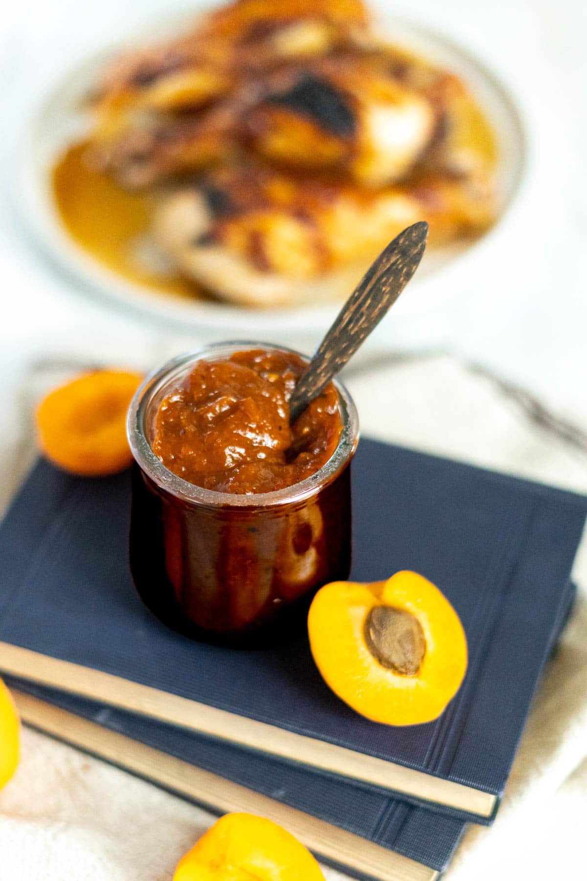 BBQ sauce in a jar with wooden spoon and sliced apricots and a plate of chicken.