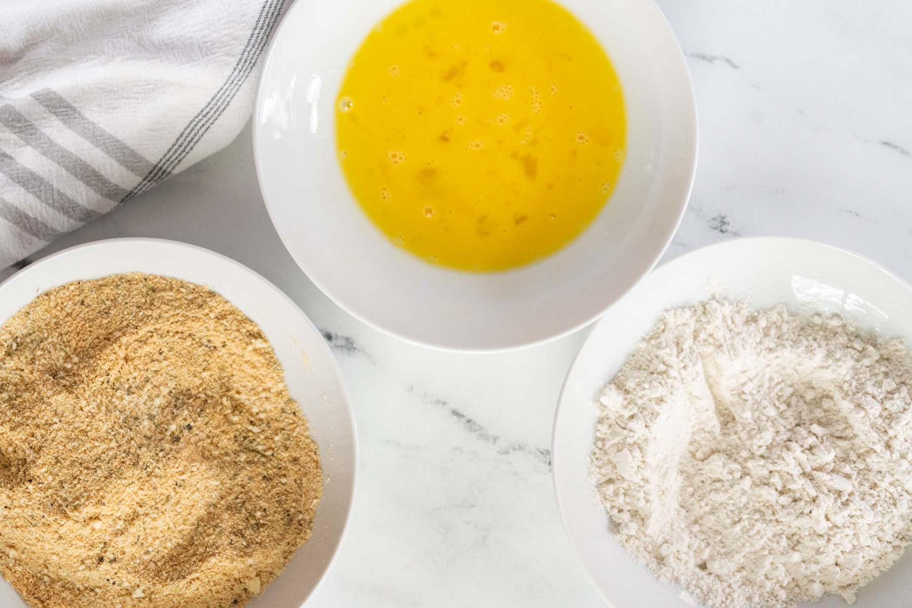 Three white bowls on a marble surface, holding the ingredients for gluten-free eggplant parmesan—beaten eggs, flour, and seasoned breadcrumbs—are arranged beside a striped kitchen towel.