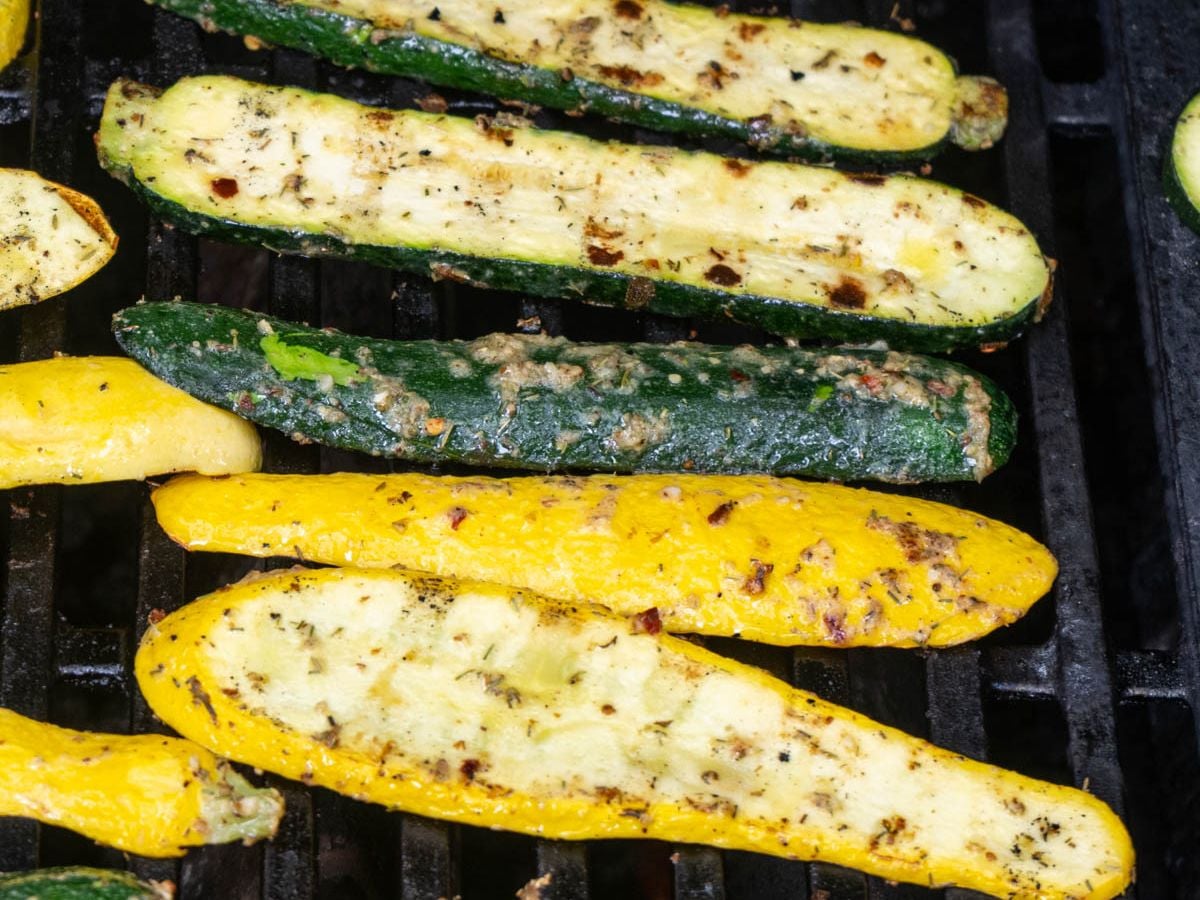 Grilled zucchini and squash halves seasoned with herbs and spices, cooked to perfection on a barbecue grill grate.
