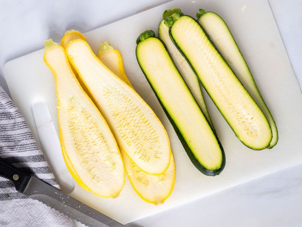 Sliced yellow squash and zucchini, perfect for grilled zucchini and squash recipes, are arranged on a white cutting board beside a knife and a striped kitchen towel.