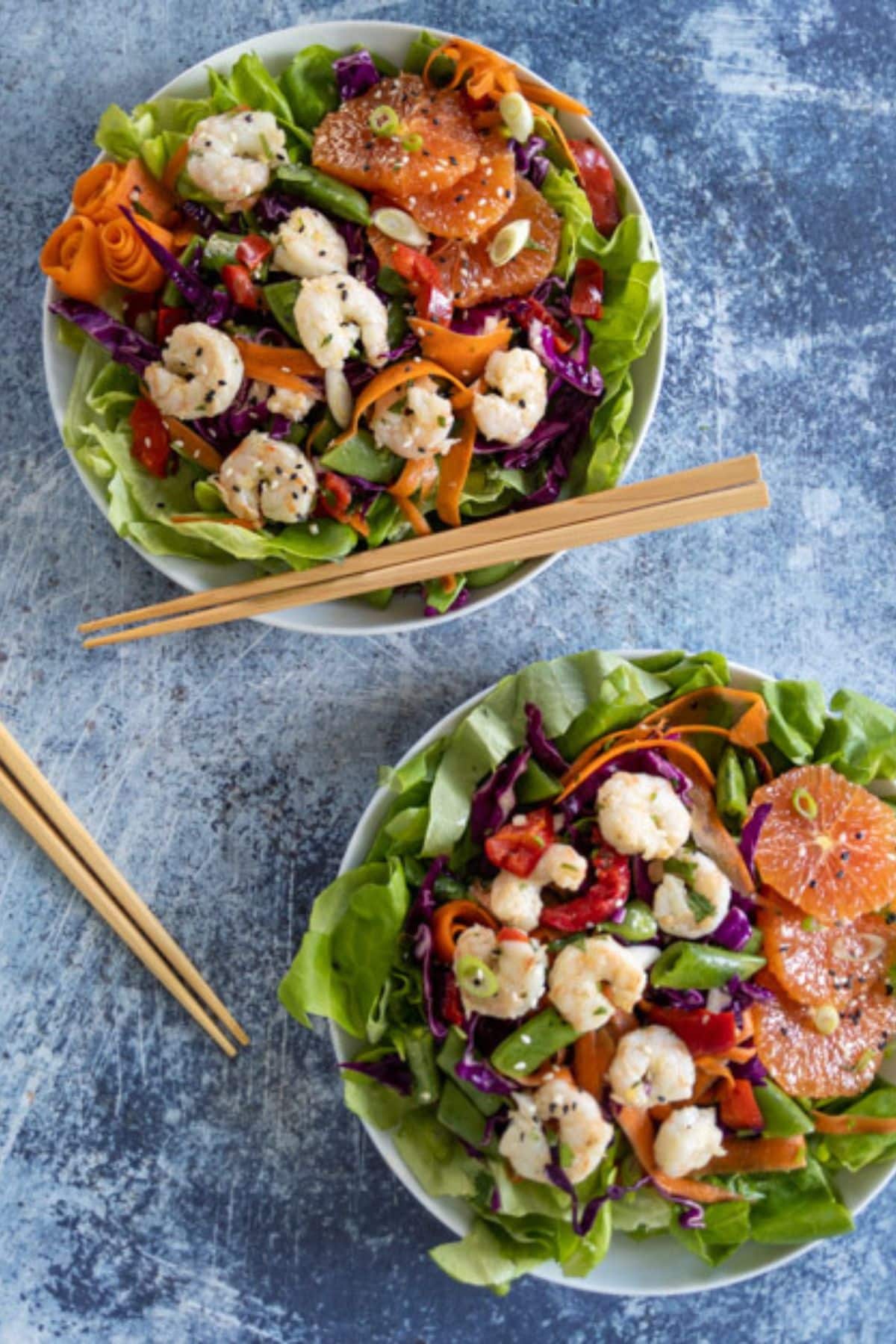 Two bowls of salad on a blue surface, featuring shrimp, lettuce, orange slices, shredded carrot, red cabbage, and green vegetables. Each bowl is paired with wooden chopsticks and drizzled with a zesty orange ginger dressing.