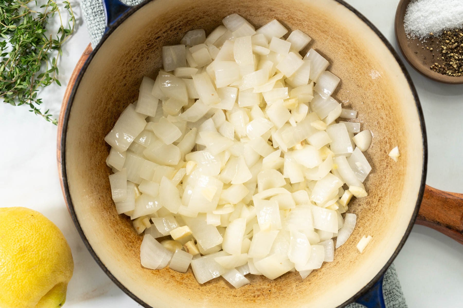 A pot filled with chopped white onions being saut&eacute;ed, with a wooden handle visible-perfect as the base for zucchini soup. Fresh thyme, a lemon, plus small bowls of salt and pepper are arranged on the surrounding white surface.