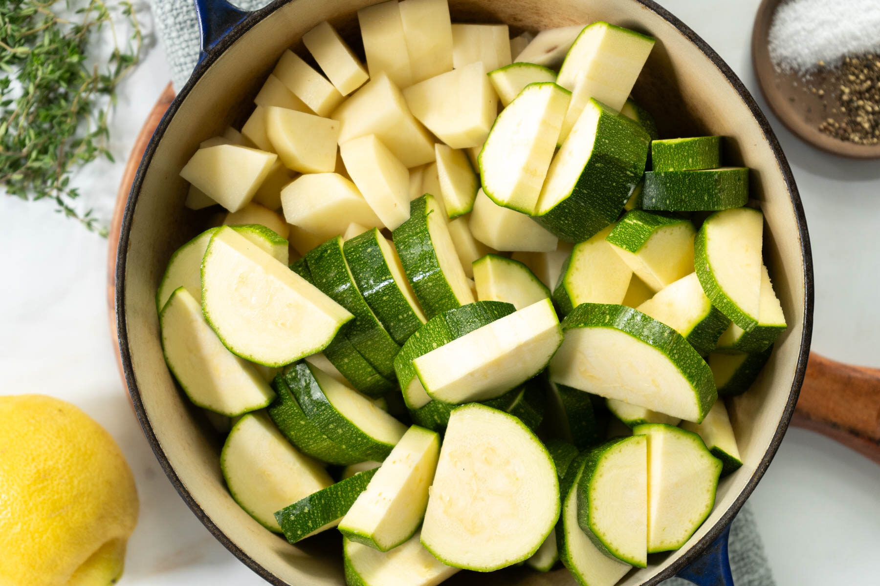 A pot filled with chopped zucchini and potatoes on a white surface, ready to be transformed into a comforting zucchini soup, with a lemon, fresh herbs, a bowl of salt, and a bowl of pepper nearby.