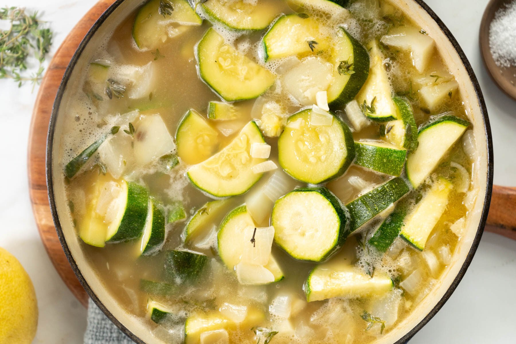 A pot of zucchini soup simmers with sliced zucchini, chopped onions, fresh herbs, and savory broth. Sprigs of herbs float atop the zucchini soup, enhancing its inviting aroma.